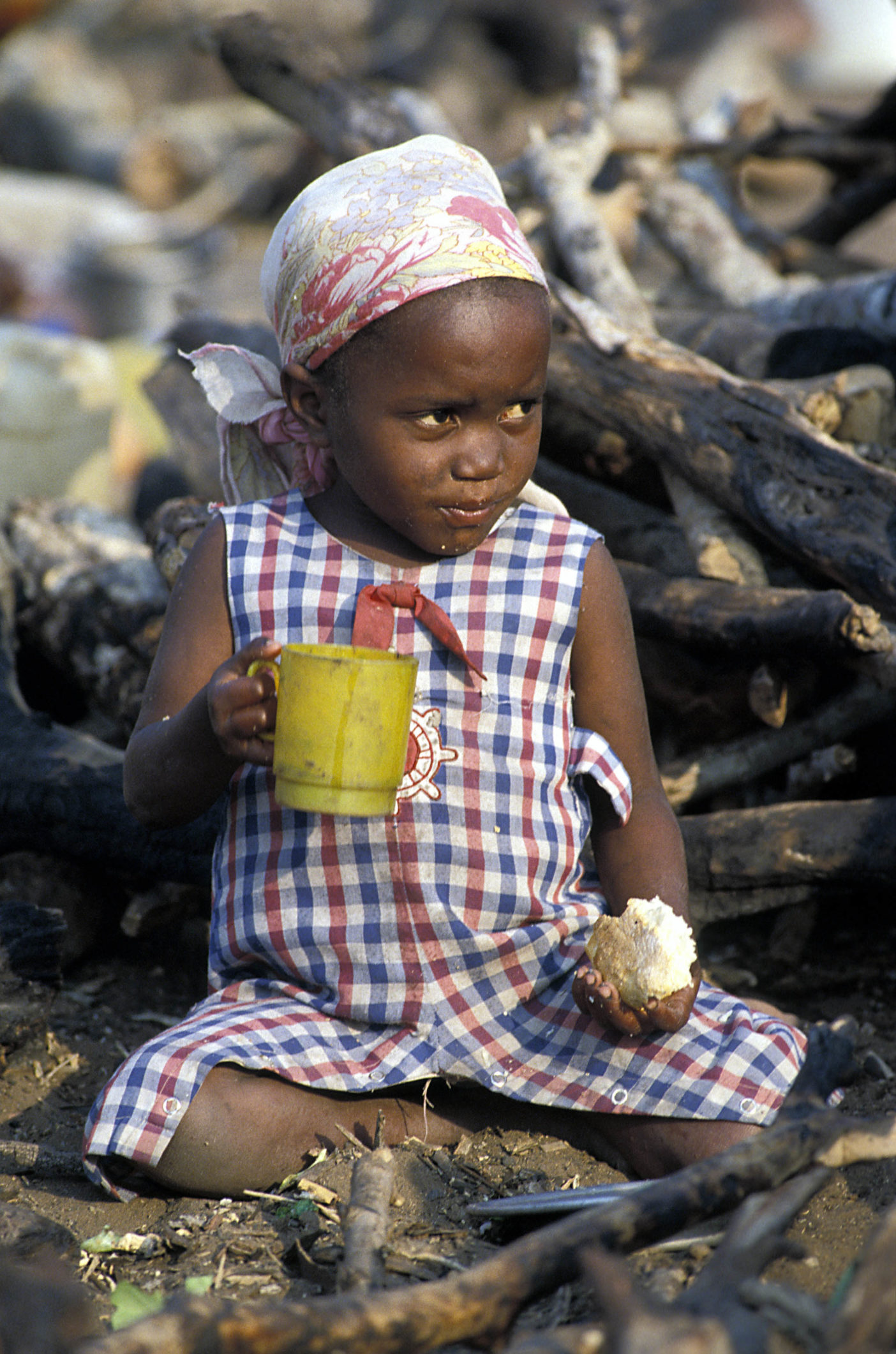 Maputo, Mozambico 1990. Bambina africana seduta per terra nella sporcizia con un tozzo di pane ed una tazza 
