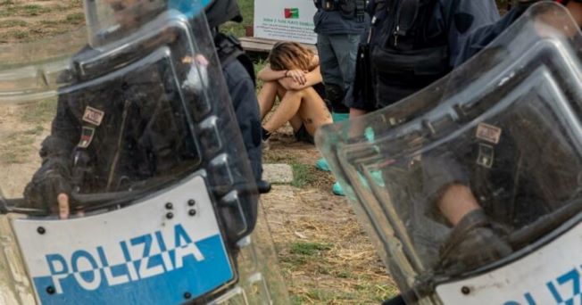 Foto: un'attivista accovacciata al rifugio Cuori Liberi, dietro gli agenti in assetto antisommossa (credits: Martina Miccichè e Saverio Nichetti)